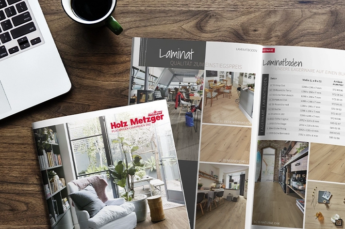 Von oben aufgenommen, zeigt die braune Holz-Tischplatte einen aufgeschlagenen Holz-Metzger-Katalog neben einer Kaffeetasse und einem aufgeklappten Laptop.