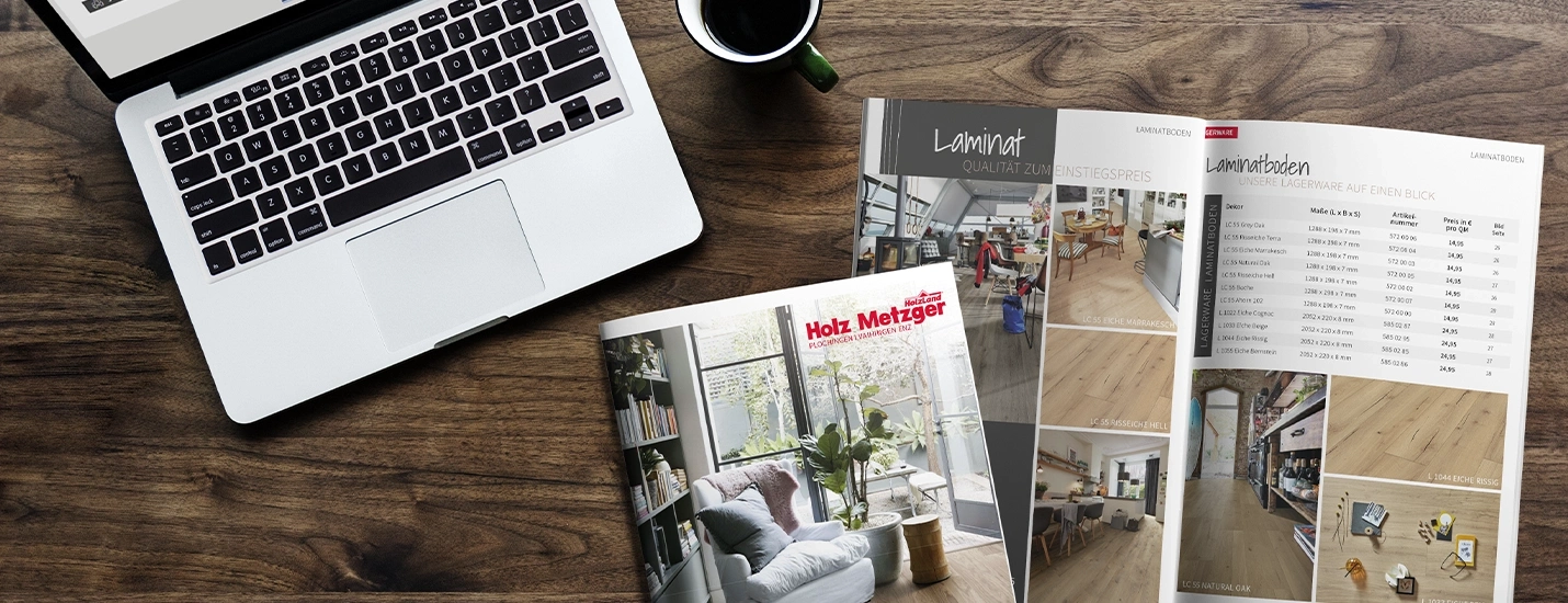 Von oben aufgenommen, zeigt die braune Holz-Tischplatte einen aufgeschlagenen Holz-Metzger-Katalog neben einer Kaffeetasse und einem aufgeklappten Laptop.