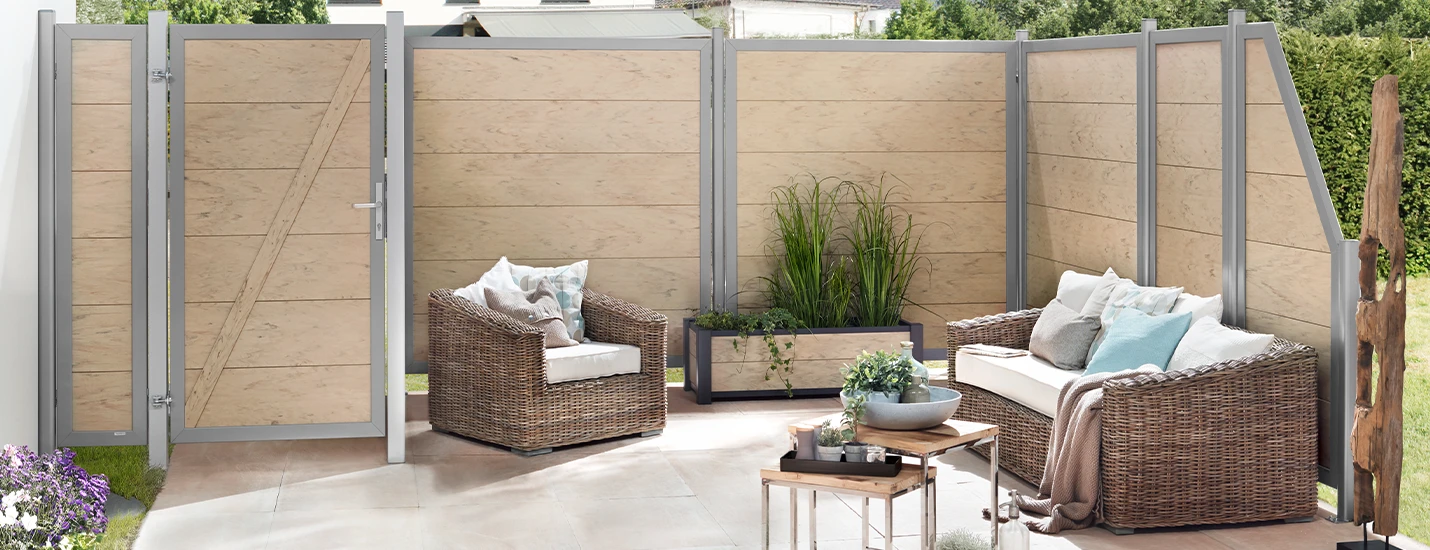 Mehrere Sichtschutzelemente und ein Eingangstor umgeben einen Bereich. Die Terrasse ist von einem Zaun eingerahmt. Auf der Terrasse stehen Gartenstühle, ein Tisch und mehrere Topfpflanzen.